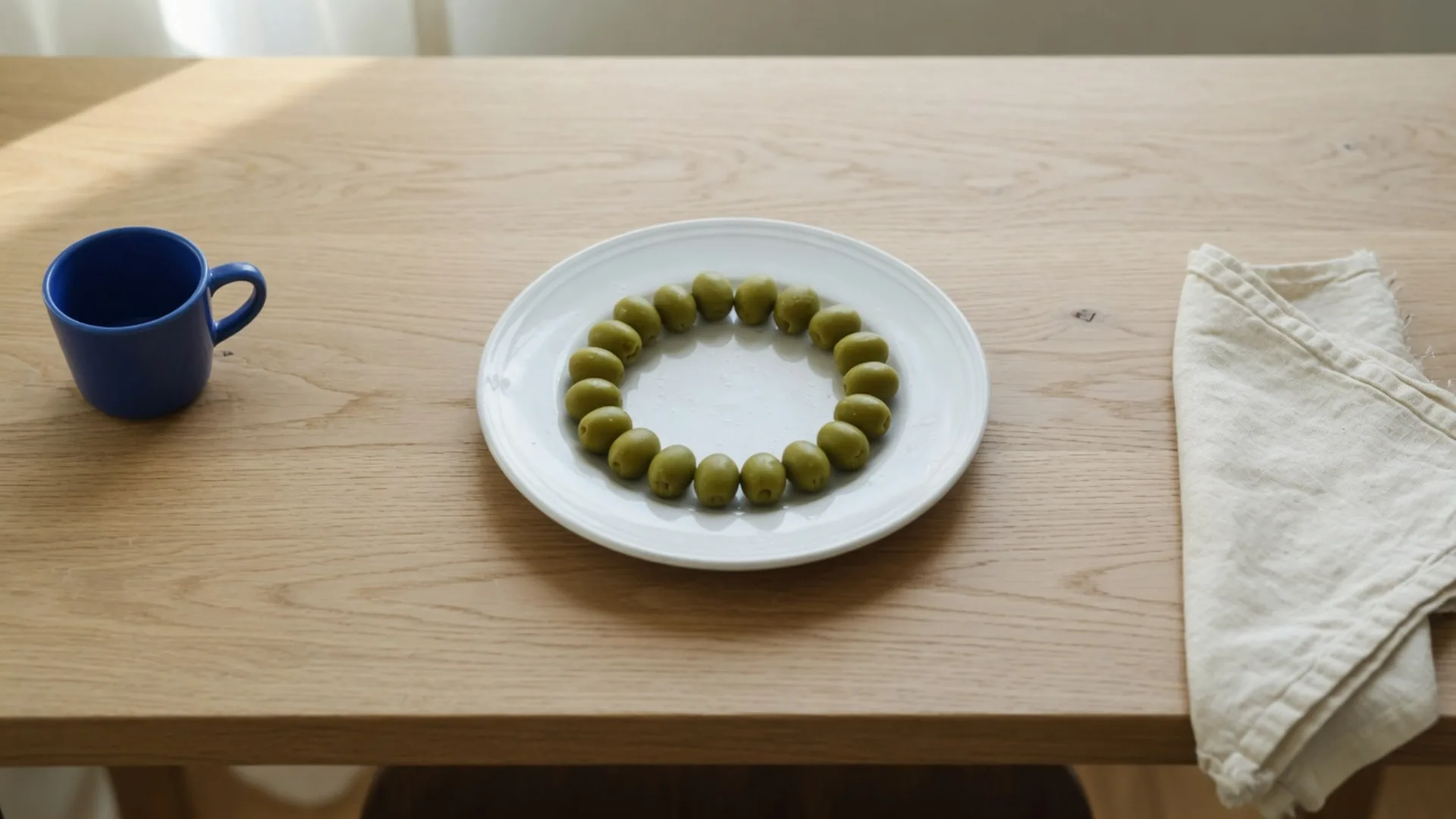 A 16:9 oak table with a round plate of olives, a cobalt cup at the left edge, and a linen napkin at the right edge